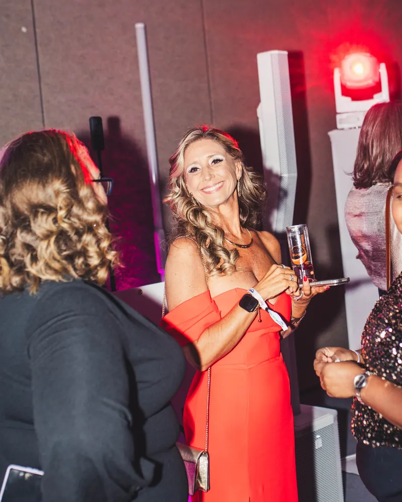 A guest in a red dress smiling with a drink beside the white DJ setup and pink lights at a holiday gala