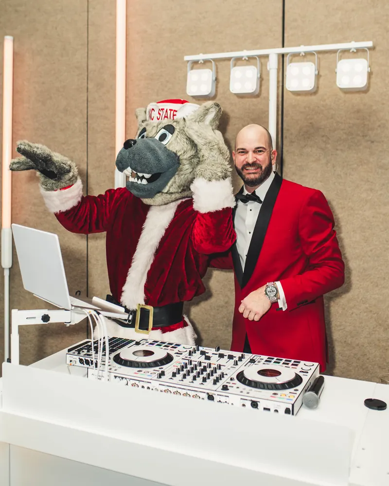 Justin at the white DJ booth with NC State mascot Mr. Wuf in a Santa suit at a holiday gala