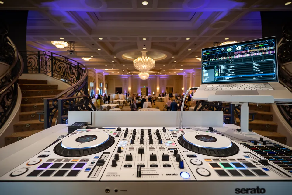 A white DJ controller and laptop at the edge of a ballroom, purple uplighting and crystal chandeliers over the guests