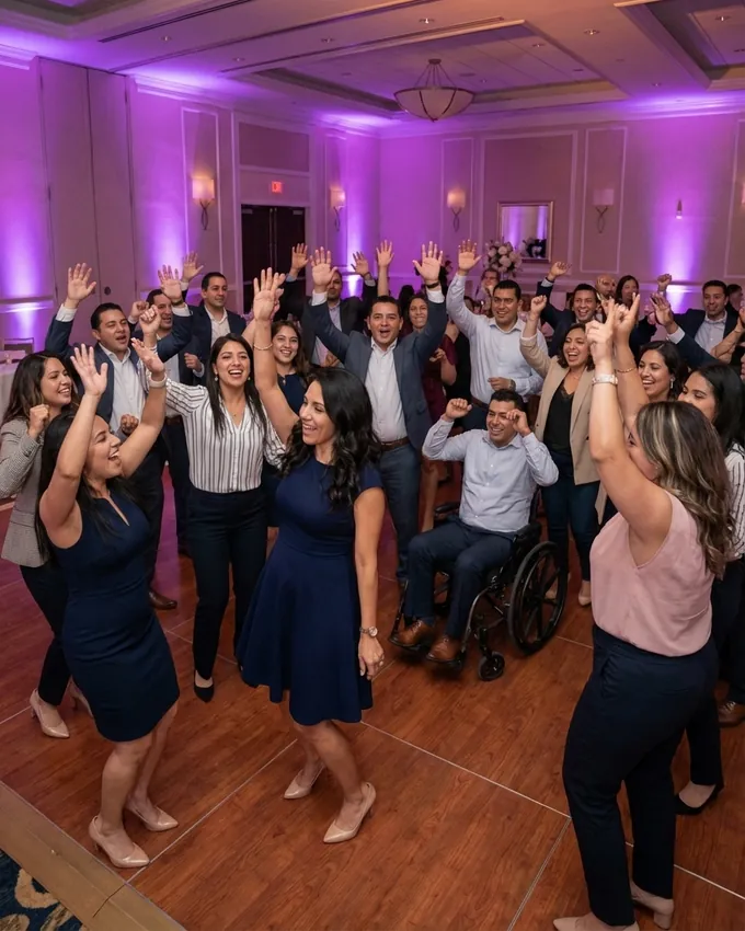 Coworkers in business clothes with their hands in the air on a packed dance floor under purple uplighting