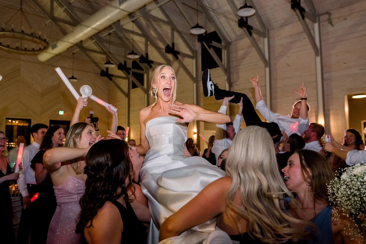 A bride lifted above a packed wedding dance floor as guests cheer and wave light up foam sticks